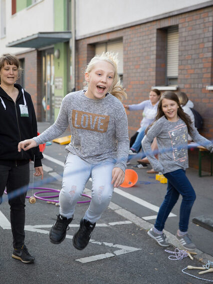 Kinder spielen auf der Straße, Erwachsene unterhalten sich. Ein Mädchen springt mit dem Seil.