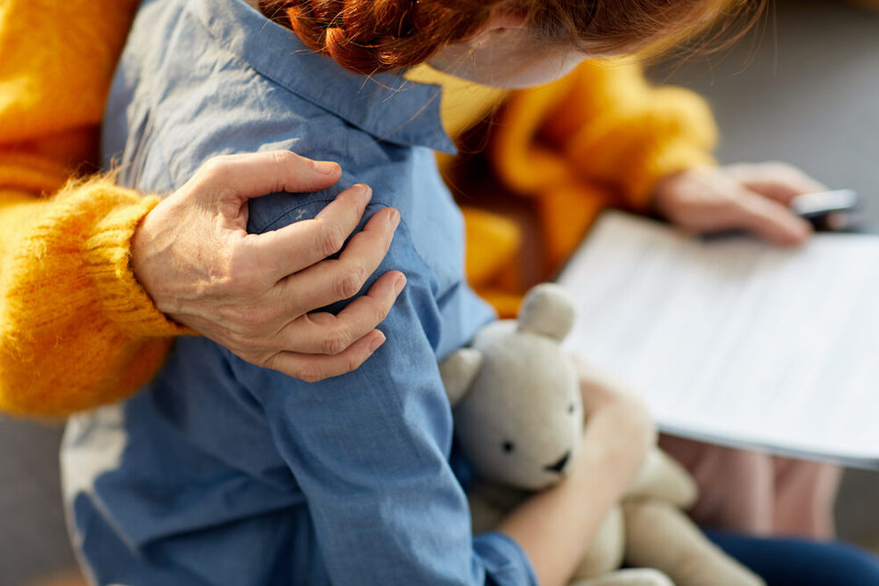 Ein Frau hält den Arm schützend um ein Mädchen. Das Kind hält einen Teddy im Arm, die Frau hält ein Formular in der Hand.