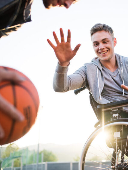 Jugendlicher im Rollstuhl spielt mit 2 Freunden draußen Basketball.