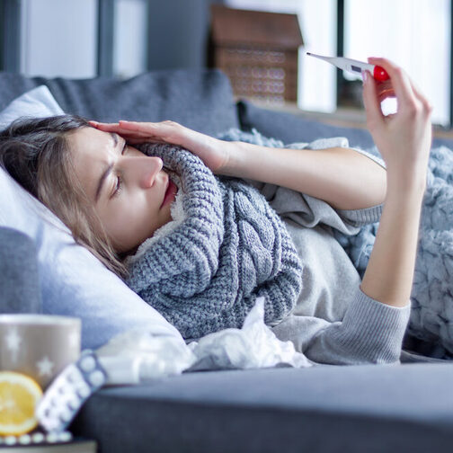 Eine Frau liegt mit einer Erkältung auf dem Sofa, eingehüllt in eine Decken, in der Hand ein Fieberthermometer