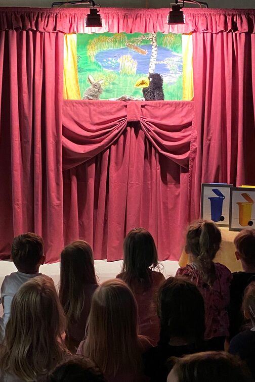 Eine Puppentheaterbühne mit rotem Vorhang. Kinder schauen beim Theaterstück mit zwei Handpuppen, einem Hasen und Raben, zu.