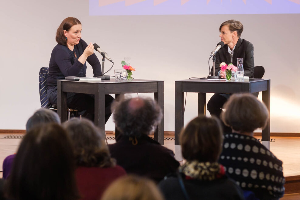 Zwei Frauen sitzen an zwei Tischen mit Mikrofonen auf einer Bühne.