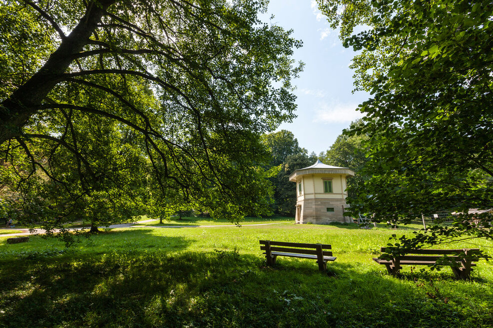 Blick auf ein kleines Steinhäuschen im Rotwildpark im Sommer. Im Vordergrund sind Sitzbänke zwischen Bäumen aufgestellt.
