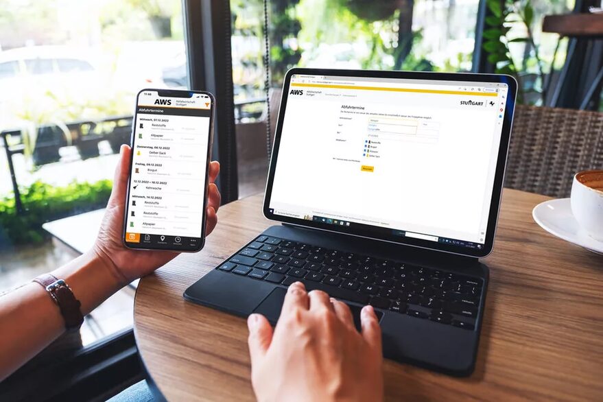 Eine Person sitzt an einem Tisch und hält ein Handy neben einen Laptop, sodass beide Bildschirme nebeneinander zu erkennen sind. Darauf ist jeweils der Online-Abfuhrkalender der AWS aufgerufen.
