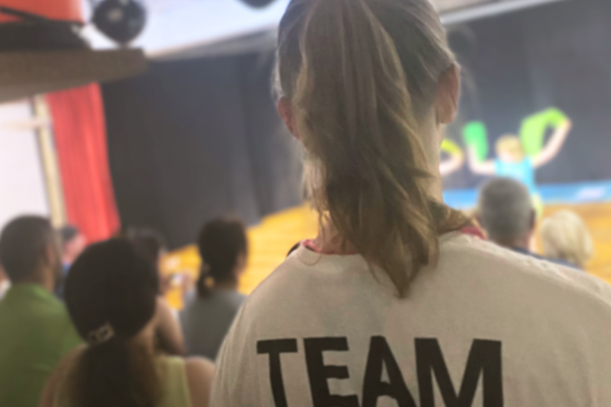 Der Rücken eines Tathea Teammitglieds mit der Aufschrift Team auf dem T-Shirt ist im Vordergrund zu sehen. Im Hintergrund ist eine Bühnenshow sowie Zuschauer*innen zu sehen.