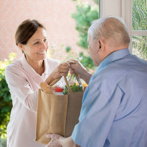 Nachbarschaftshilfe: Eine Frau überreicht einem älteren Mann eine Einkaufstasche an der Haustür.