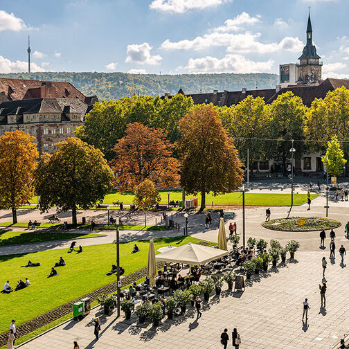 Auf dem Bild ist der Schlossplatz Stuttgart mit dem Alten Schloss zu sehen. Das Wetter ist schön, die Sonne scheint. Zu sehen sind Menschen, die flanieren.