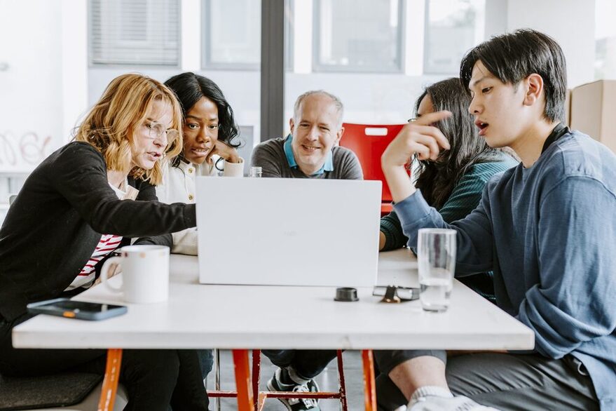 Zwei Mänenr und eine Frau sitzen an einem Tisch. Die Frau in der Mitte macht sich Notizen.