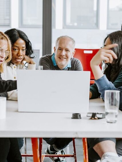 Zwei Mänenr und eine Frau sitzen an einem Tisch. Die Frau in der Mitte macht sich Notizen.