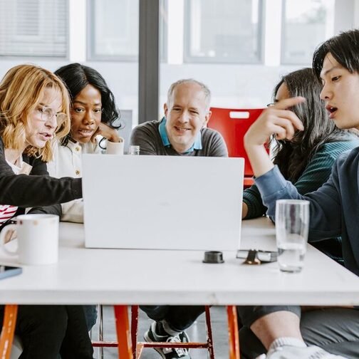 Zwei Mänenr und eine Frau sitzen an einem Tisch. Die Frau in der Mitte macht sich Notizen.
