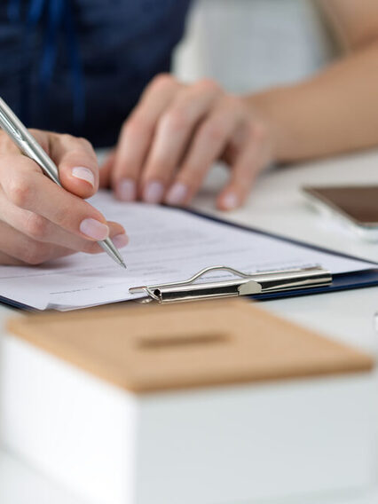 Die Hände einer Frau am Schreibtisch, die einen Bogen auf einem Klemmbrett ausfüllt.