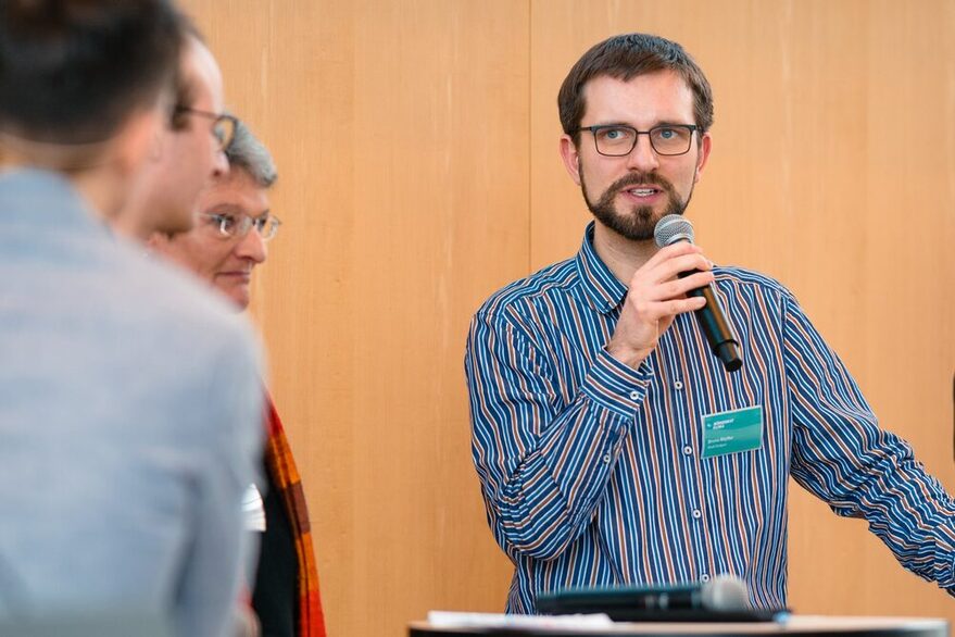 An einem Stehtisch steht ein Mann mit einem Mikrofon in der Hand. Neben ihm stehen drei weitere Personen.