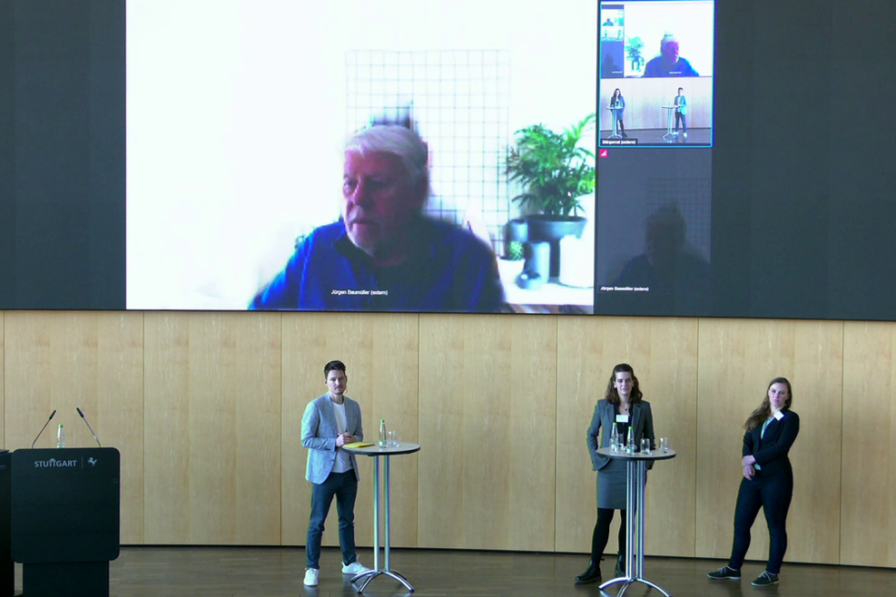 Drei Menschen stehen auf der Bühne, ein vierter ist per Video auf der Leinwand dahinter