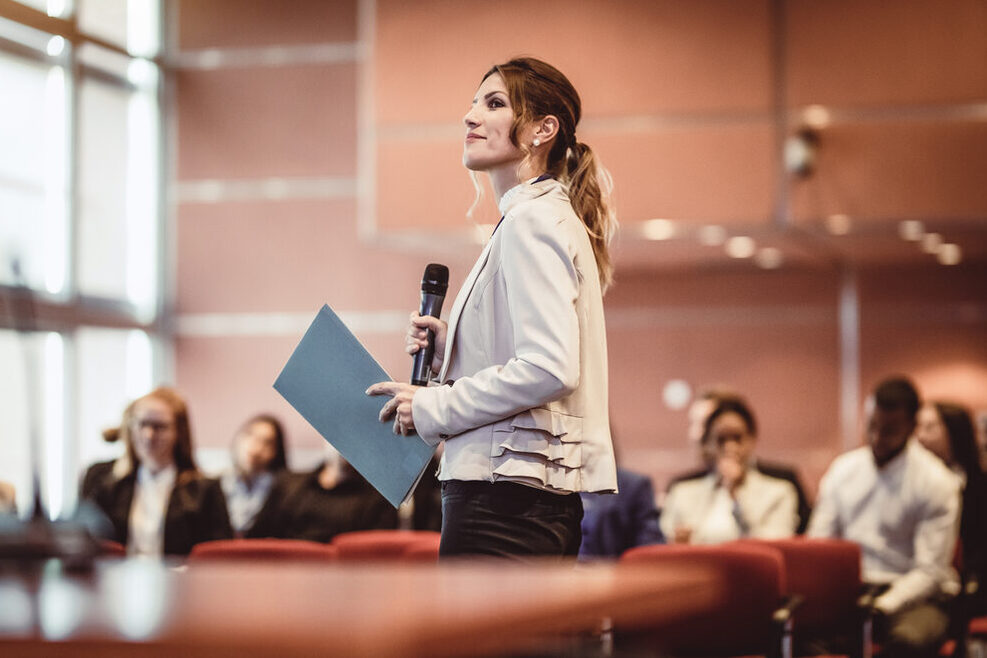Eine Frau steht mit einem Mikrofon der Hand vor einem Publikum