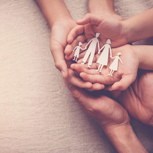 Ein Erwachsener und ein Kind halten die Hände zusammen. In den Kinderhänden liegt ein Scherenschnitt einer vierköpfigen Familie.