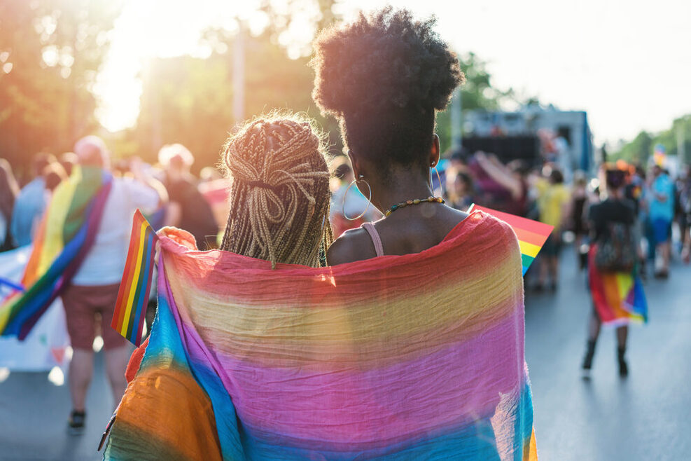 Zwei Frauen stehen Arm in Arm auf einer Veranstaltung im Freien, um die Schulter tragen Sie eine Regenbogenfahne.