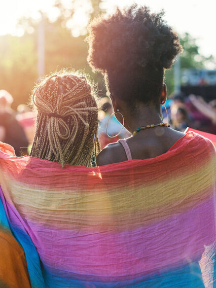 Zwei Frauen stehen Arm in Arm auf einer Veranstaltung im Freien, um die Schulter tragen Sie eine Regenbogenfahne.