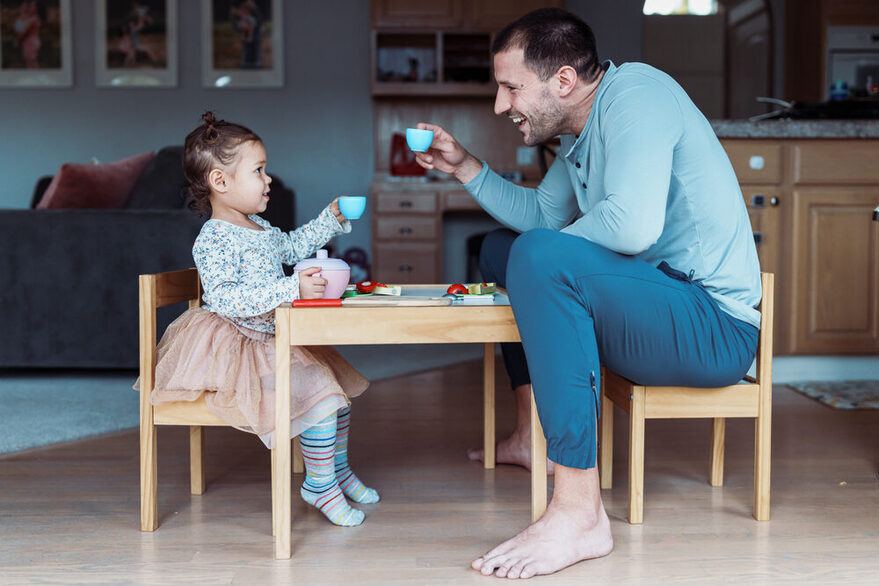 Ein Kleinkind spielt Tee-trinken mit seinem Vater in einem Wohnzimmer an einem Spieltisch.