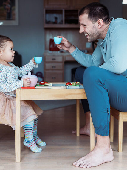 Ein Kleinkind spielt Tee-trinken mit seinem Vater in einem Wohnzimmer an einem Spieltisch.