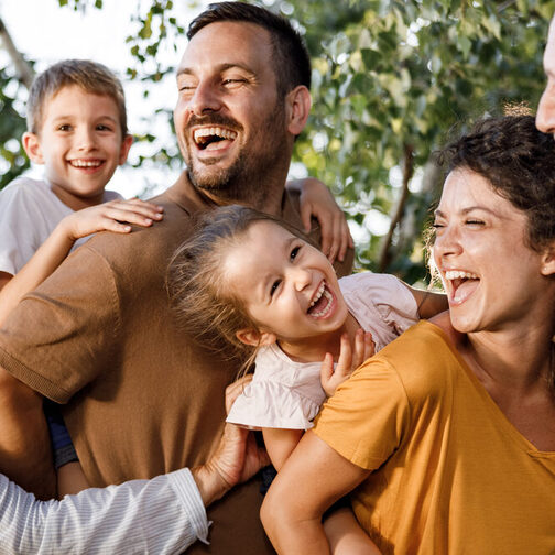 Familie mitsamt Großeltern, Vater, Mutter, Tochter und Sohn lachen spielerisch in der Natur.