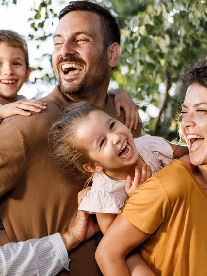 Familie mitsamt Großeltern, Vater, Mutter, Tochter und Sohn lachen spielerisch in der Natur.