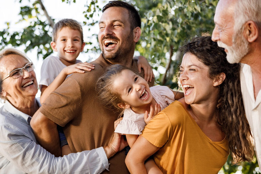Familie mitsamt Großeltern, Vater, Mutter, Tochter und Sohn lachen spielerisch in der Natur.