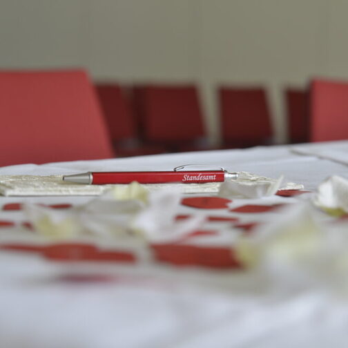 Ein roter Kugelschreiber mit dem Aufdruck Standesamt liegt auf einem dekorierten Tisch mit weißem Tischtuch.