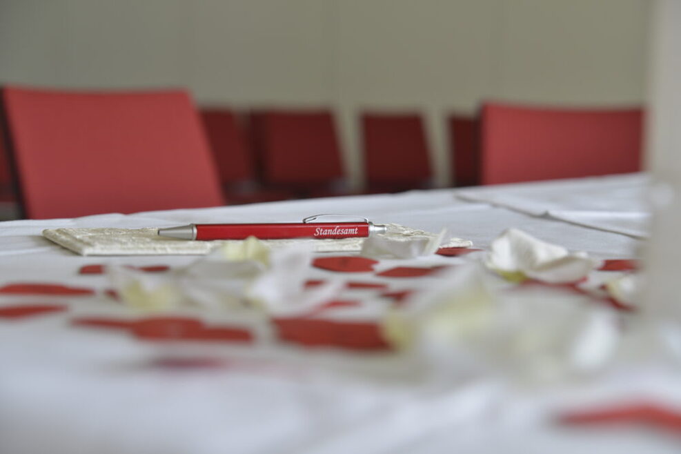 Ein roter Kugelschreiber mit dem Aufdruck Standesamt liegt auf einem dekorierten Tisch mit weißem Tischtuch.