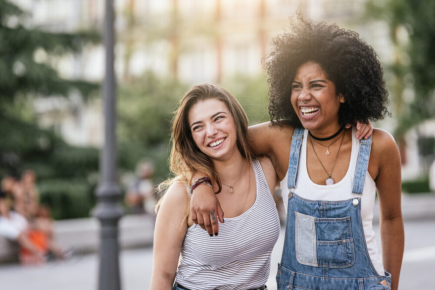 Zwei lachende Frauen stehen Nebeneinander und liegen sich in den Armen.