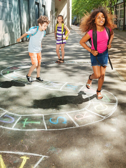 Drei Kinder hüpfen auf dem Weg zur Schule über ein Kreidbild auf der Straße.