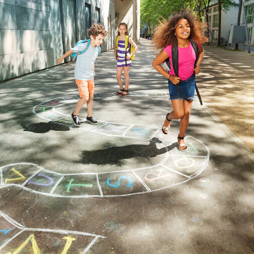 Drei Kinder hüpfen auf dem Weg zur Schule über ein Kreidbild auf der Straße.