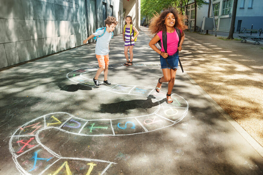 Drei Kinder hüpfen auf dem Weg zur Schule über ein Kreidbild auf der Straße.