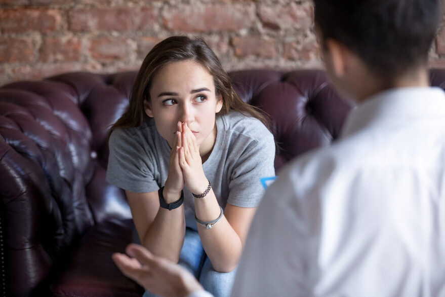 Junge Frau sitzt auf einer Couch vor einem Therapeuten. Sie blickt unglücklich in die Ferne.