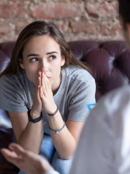 Junge Frau sitzt auf einer Couch vor einem Therapeuten. Sie blickt unglücklich in die Ferne.