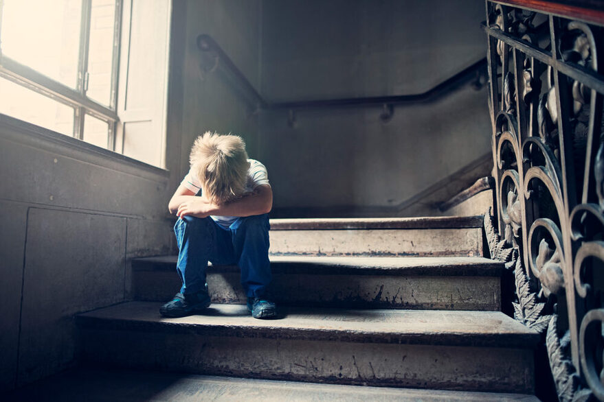 Ein kleines Kind sitzt einsam und betrübt auf einer Treppe. Es hat seinen Kopf zwischen die angezogenen Beine gesenkt.