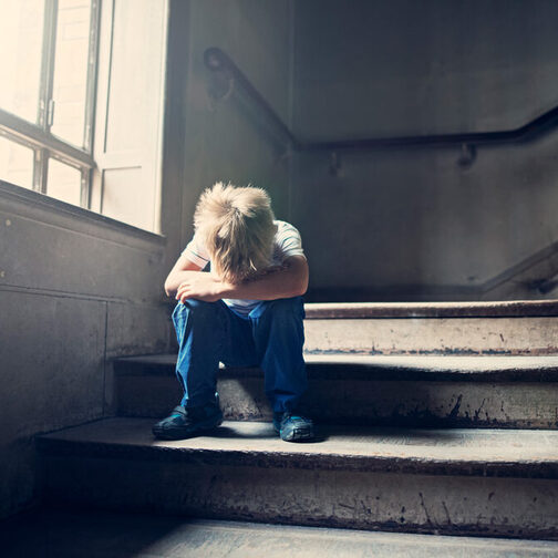 A small child sits lonely and sad on a staircase. It has lowered its head between its tucked-up legs.