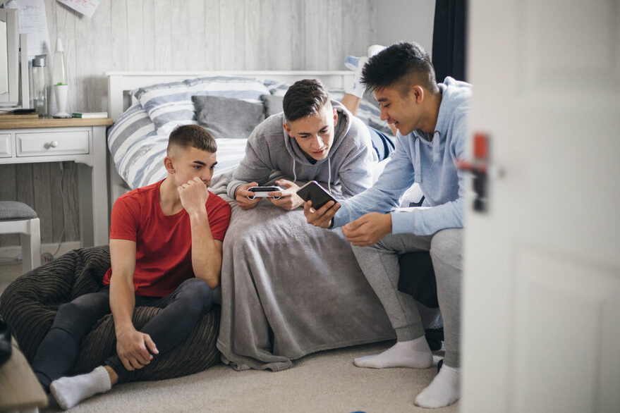 Three male teenagers sit on a bed and look at a smartphone.