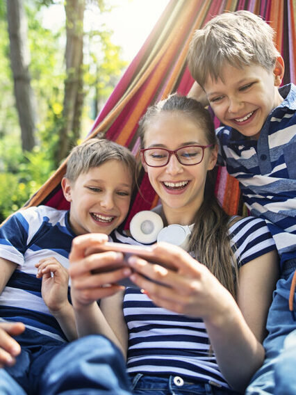 Zwei jungs und ein Mädchen liegen zu dritt in einer Hängematte und schauen lachend auf ein Smartphone.