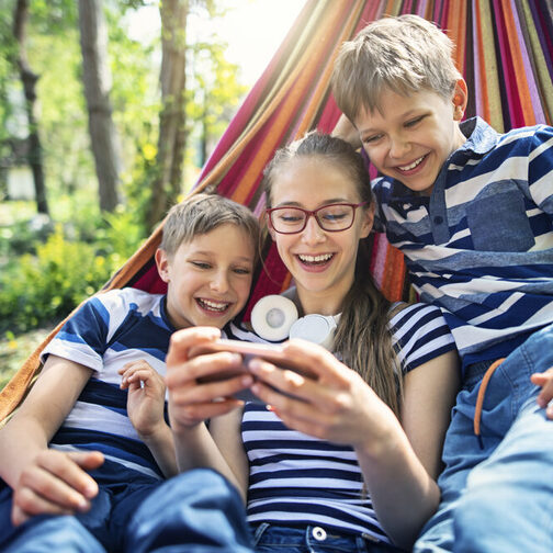 Zwei jungs und ein Mädchen liegen zu dritt in einer Hängematte und schauen lachend auf ein Smartphone.
