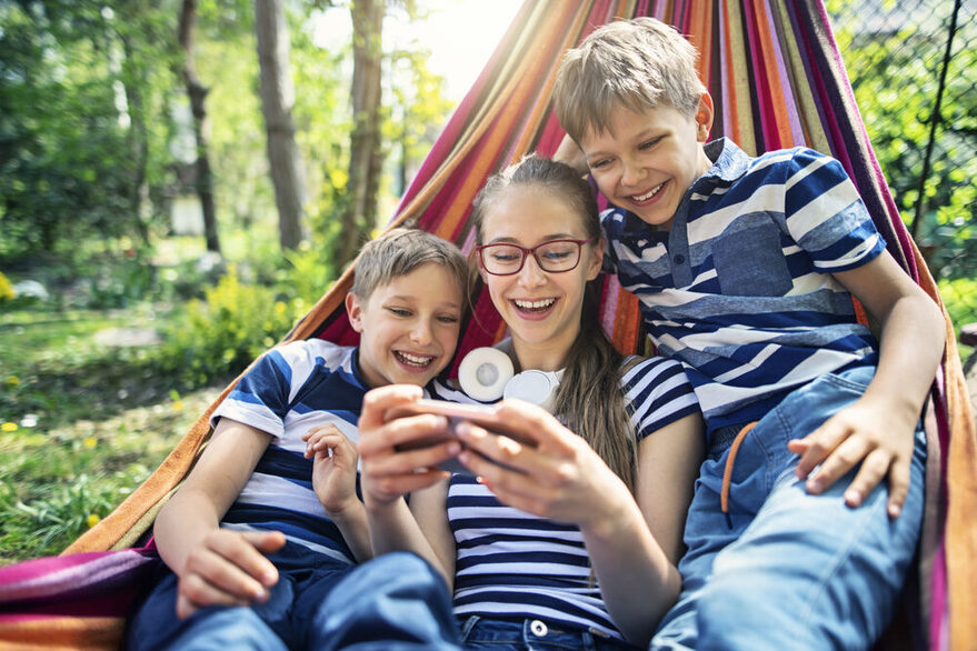 Two boys and a girl are lying in a hammock, laughing and looking at a smartphone.