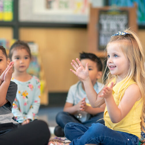 Mehrere Kinder sitzen auf dem Boden und klatschen zusammen mit einer Erzieherin.