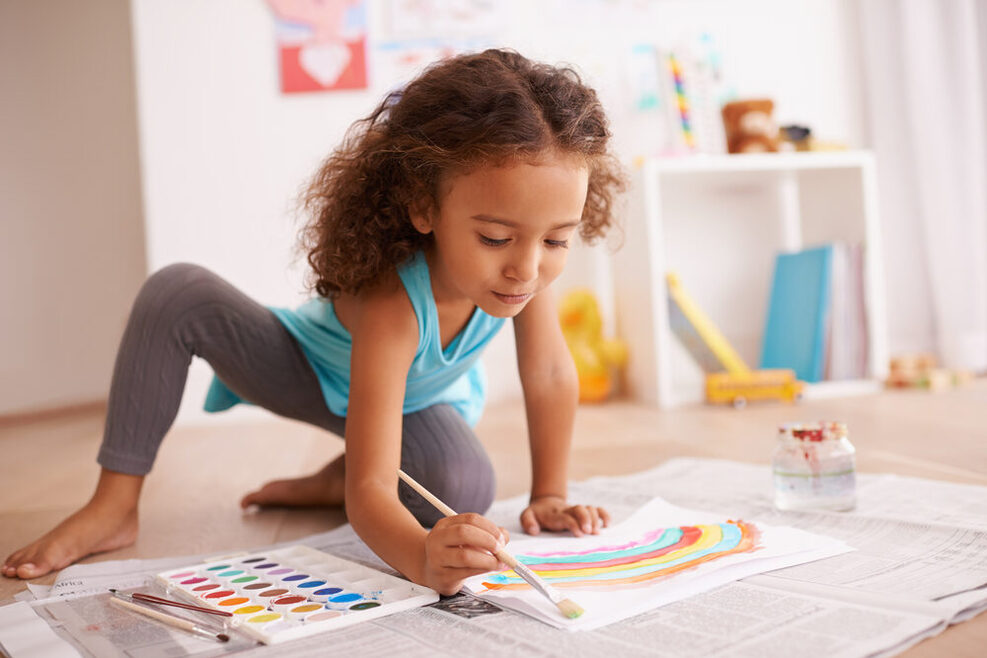 Ein Mädchen sitzt auf dem Boden und malt mit Wasserfarben einen Regenbogen auf ein Papier.