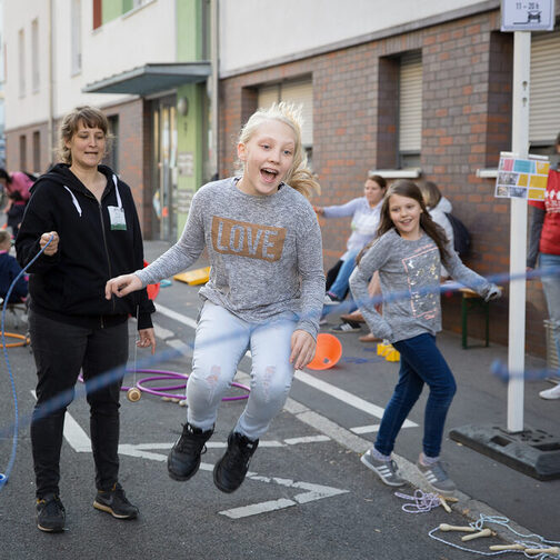 Kinder spielen auf der Straße, Erwachsene unterhalten sich. Ein Mädchen springt mit dem Seil.