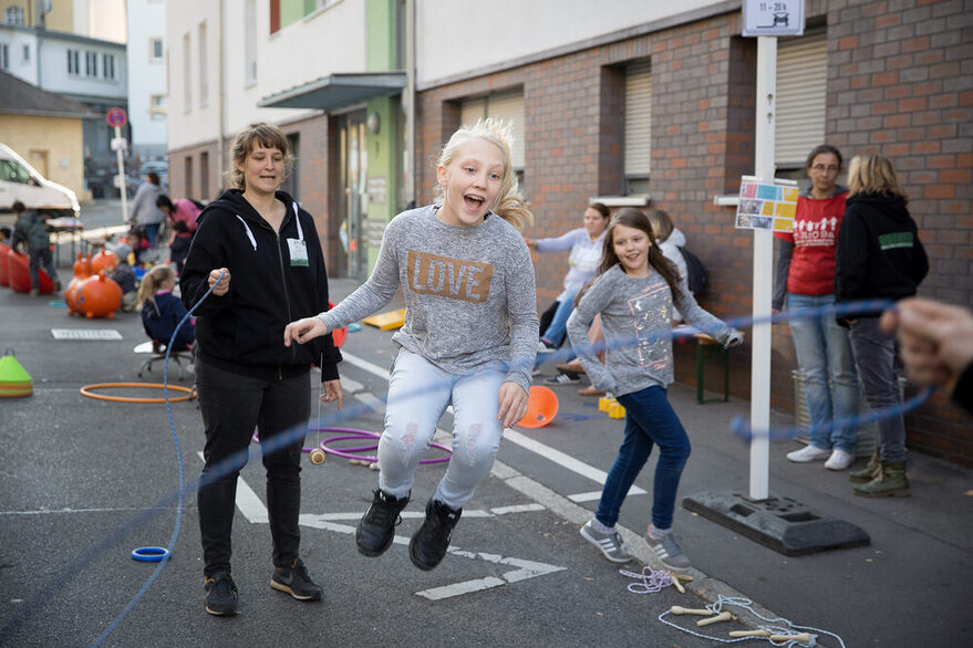 Kinder spielen auf der Straße, Erwachsene unterhalten sich. Ein Mädchen springt mit dem Seil.
