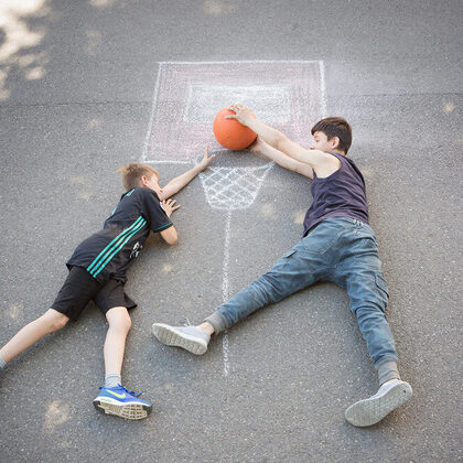 Zwei Jungen liegen auf der Straße und legen einen Ball in einen mit Kreide aufgezeichneten Basketballkorb.