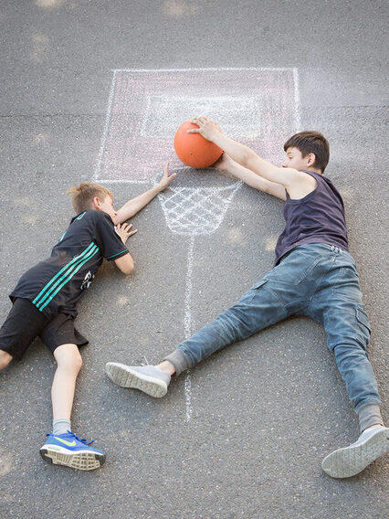 Zwei Jungen liegen auf der Straße und legen einen Ball in einen mit Kreide aufgezeichneten Basketballkorb.