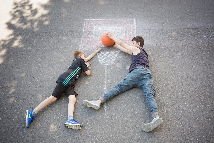 Zwei Jungen liegen auf der Straße und legen einen Ball in einen mit Kreide aufgezeichneten Basketballkorb.