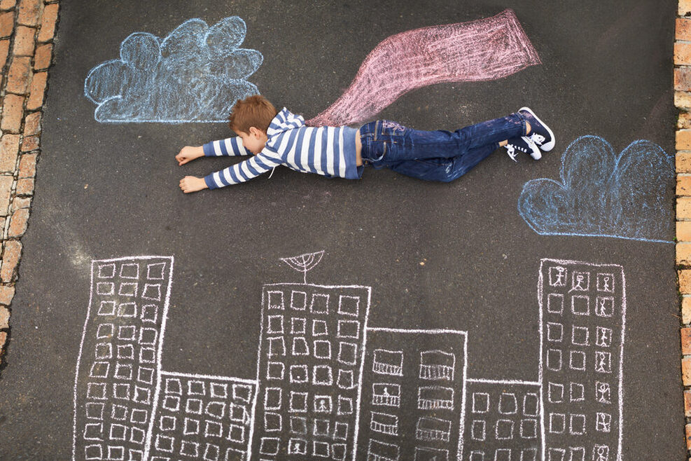 Ein Junge liegt auf einem Kreidebild auf einer Straße. Die Zeichnung lässt ihn als Superman über die Stadt fliegen.