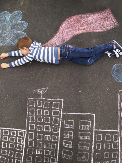 Ein Junge liegt auf einem Kreidebild auf einer Straße. Die Zeichnung lässt ihn als Superman über die Stadt fliegen.