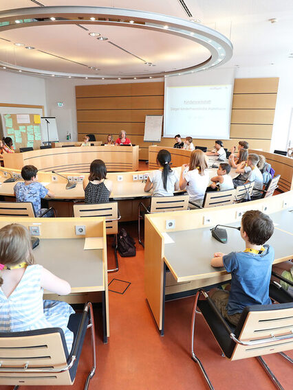 Mädchen und Jungen sitzen um runde Sitzungstische im Sitzungsraum.